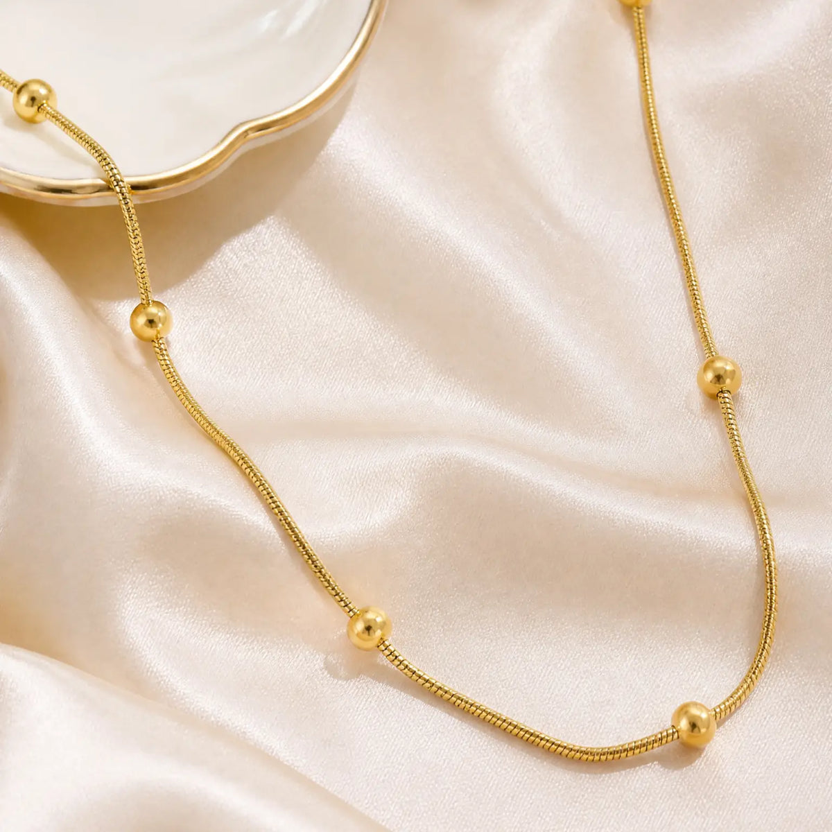 A gold chain necklace with evenly spaced round gold beads, draped elegantly on a shimmering champagne satin fabric.