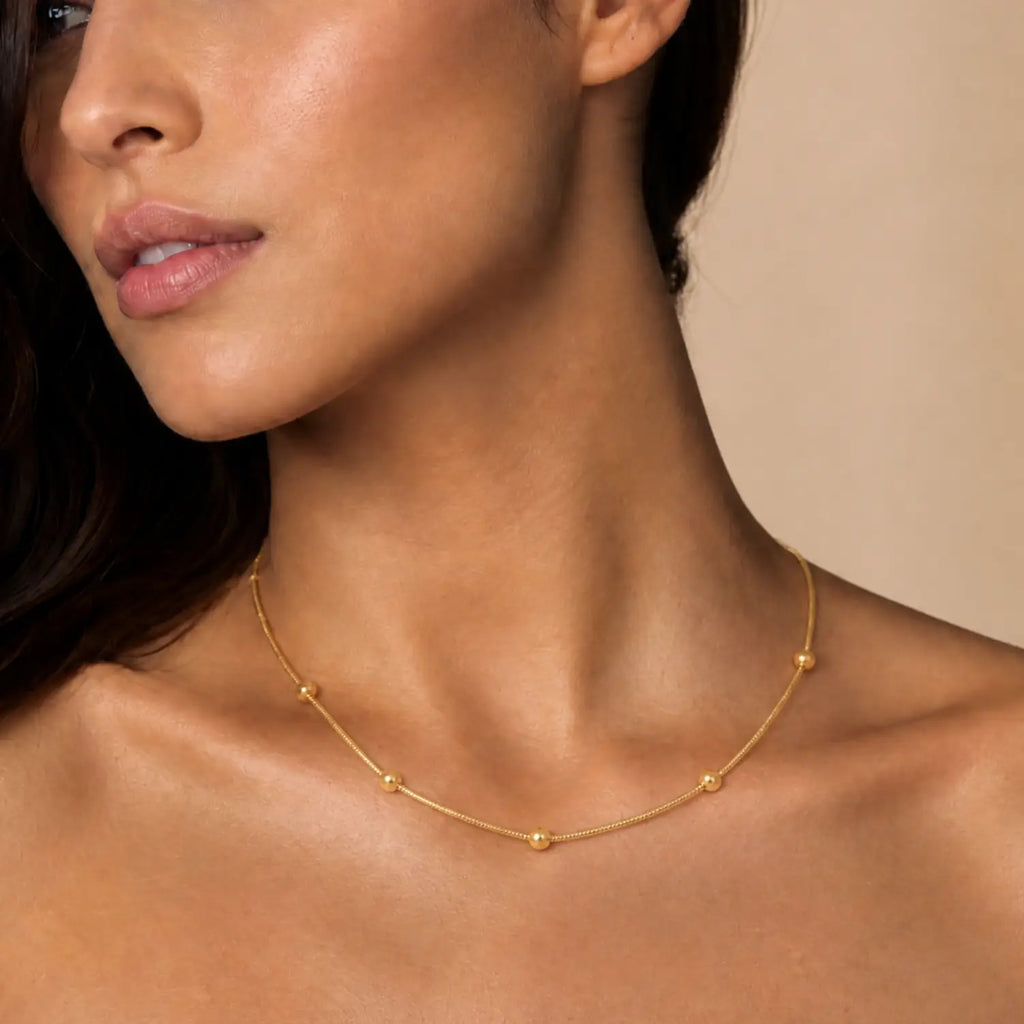 A close-up of a model wearing a delicate gold chain necklace with small spaced bead details, set against a soft beige background.
