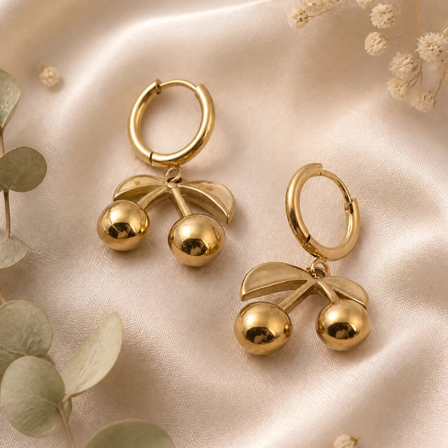 A pair of gold hoop earrings with dangling cherry motifs, displayed on a soft beige satin fabric with decorative eucalyptus leaves and dried flowers.