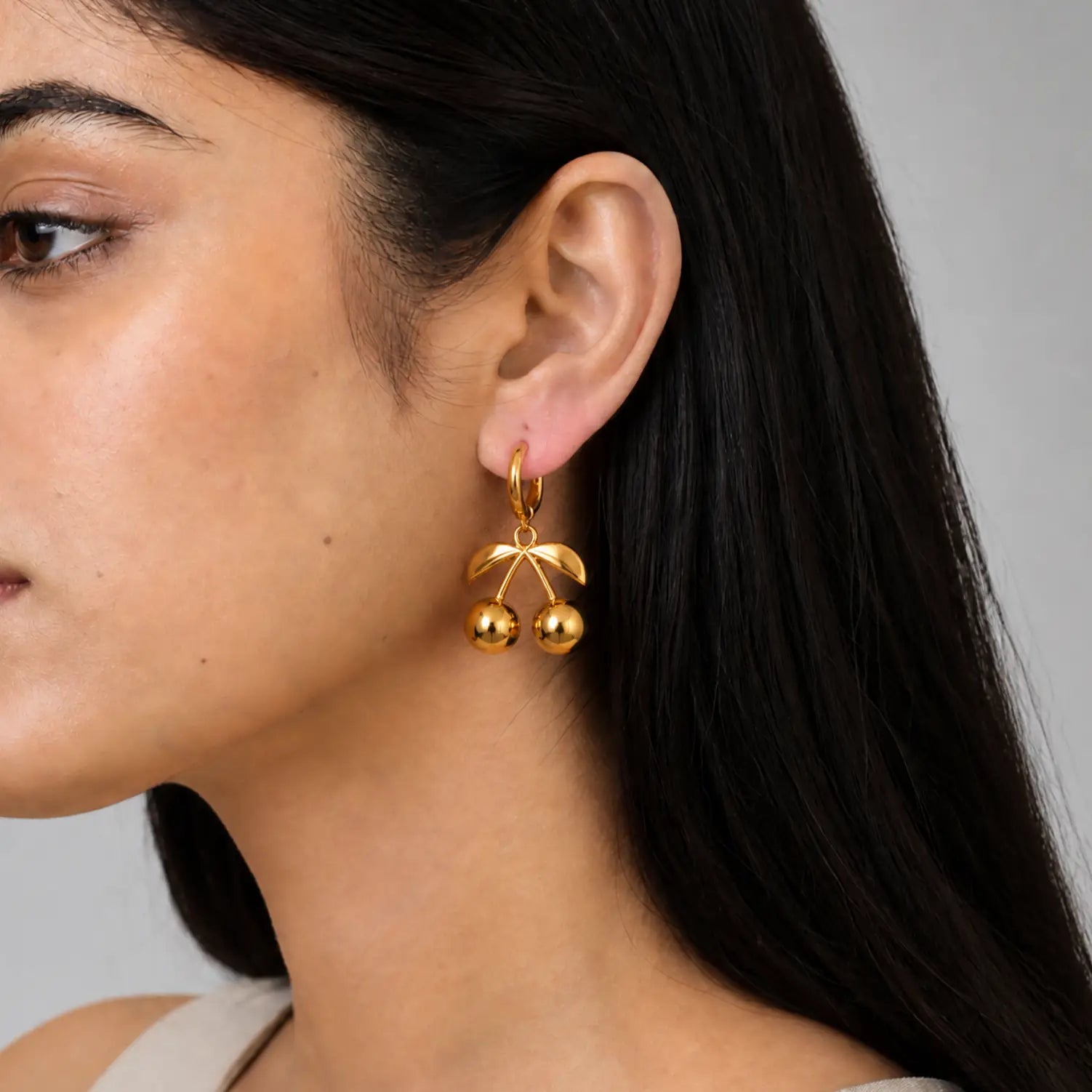 A close-up of a model wearing a gold huggie hoop earring with a dangling cherry charm featuring two small gold balls.