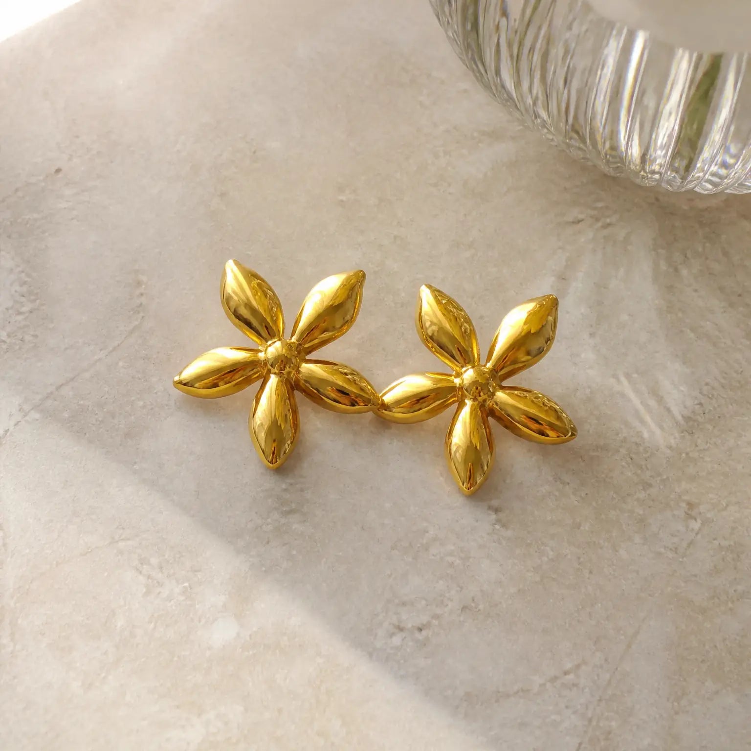 A pair of shiny gold flower-shaped stud earrings resting on a light beige textured surface, with a blurred ribbed glass object in the upper right corner.