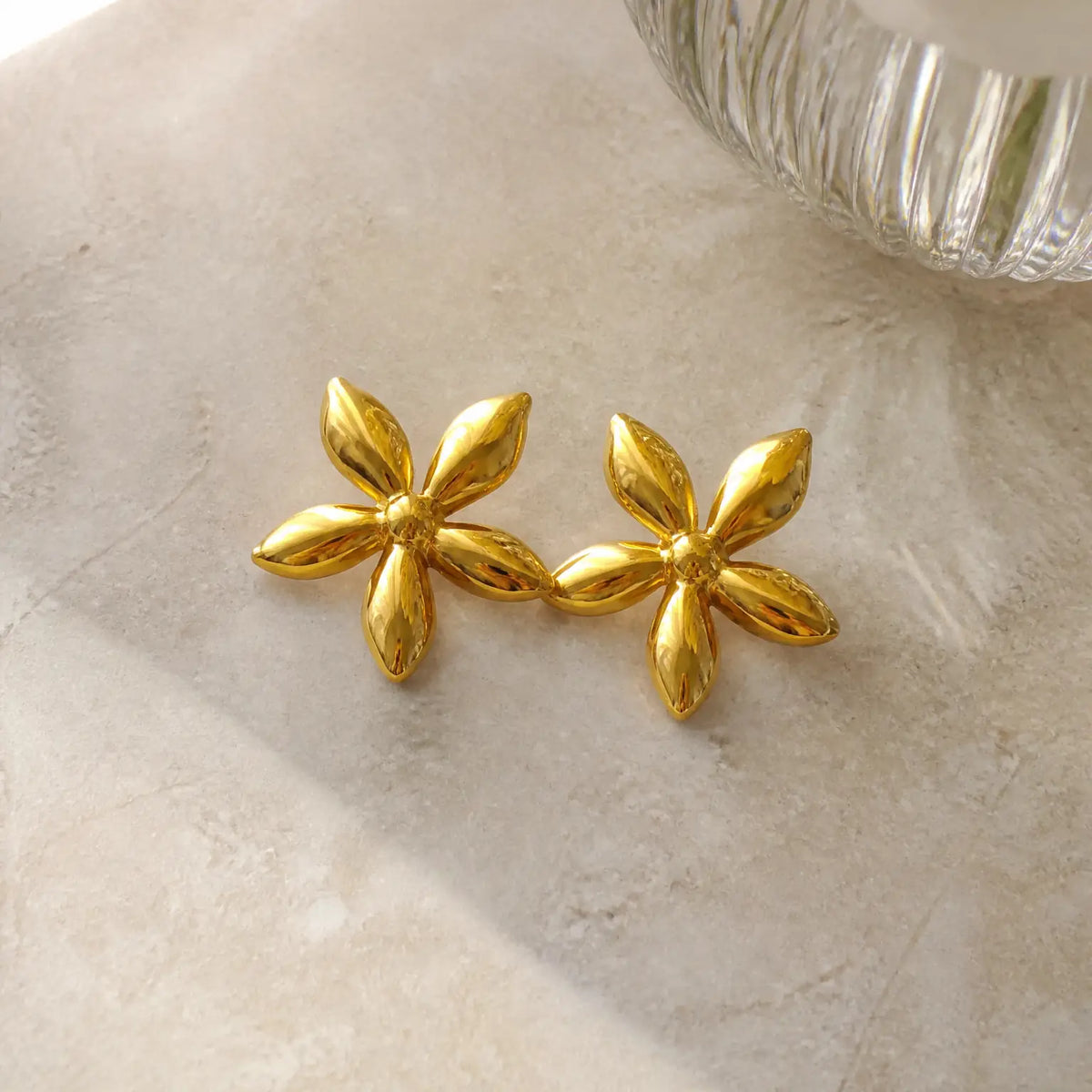 A pair of shiny gold flower-shaped stud earrings resting on a light beige textured surface, with a blurred ribbed glass object in the upper right corner.