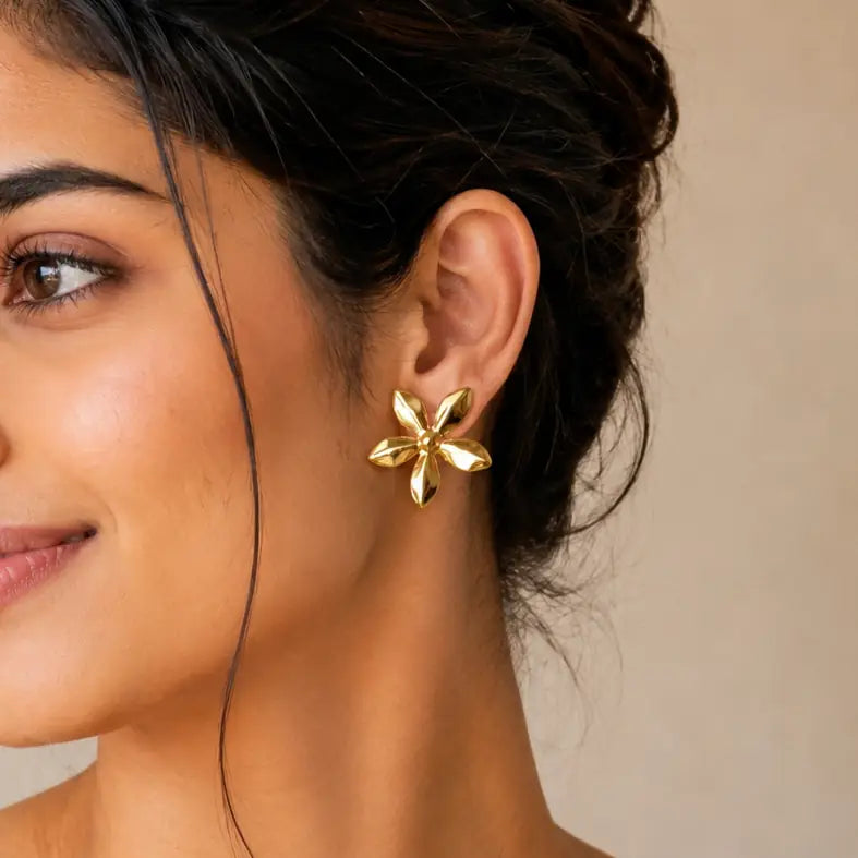 A close-up of a woman's ear wearing a gold flower-shaped stud earring against a soft beige background.