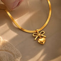 A gold snake chain necklace with a bow and heart pendant, styled on a beige surface with warm sunlight and shadows.