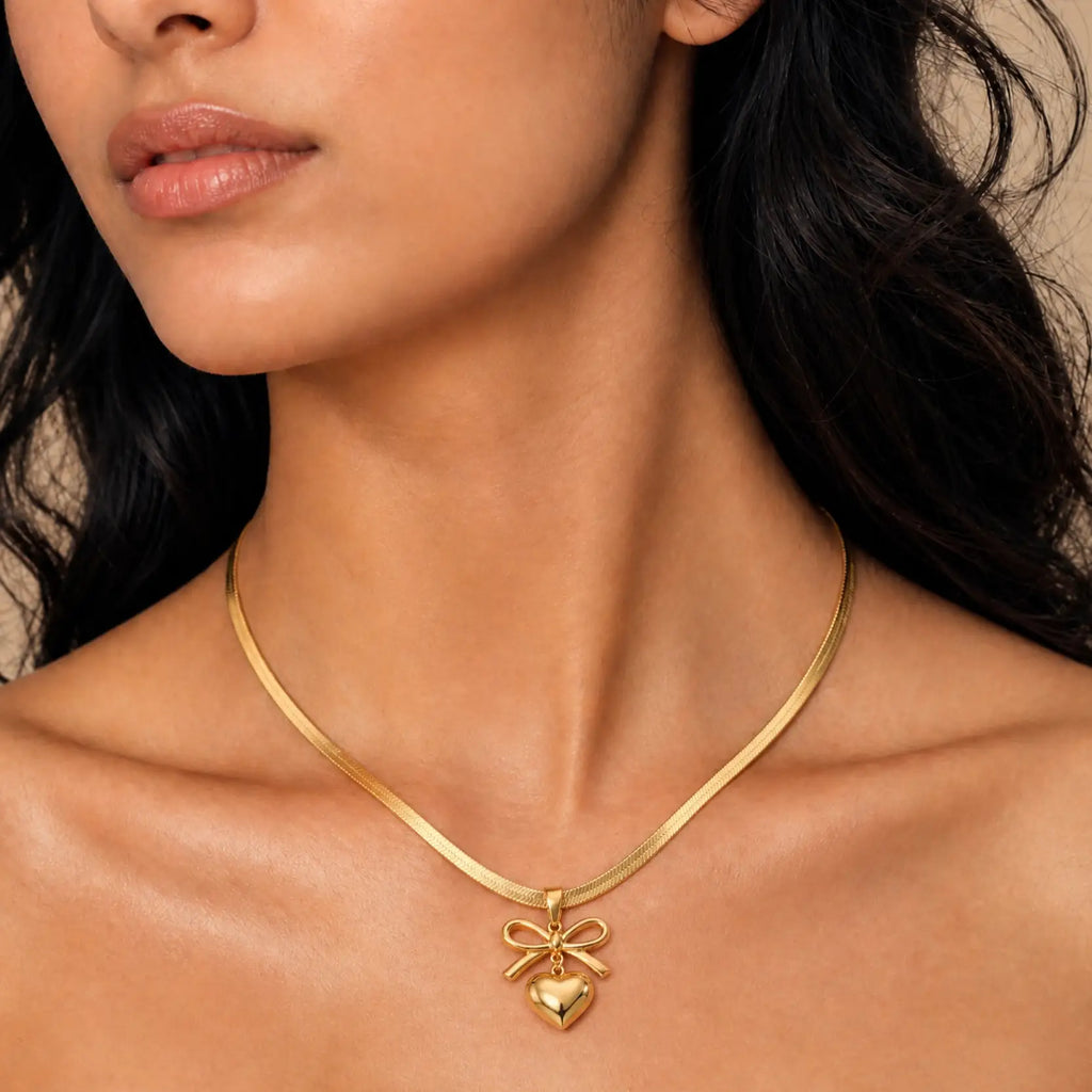 A close-up of a model wearing a delicate gold snake chain necklace with a bow and heart pendant.