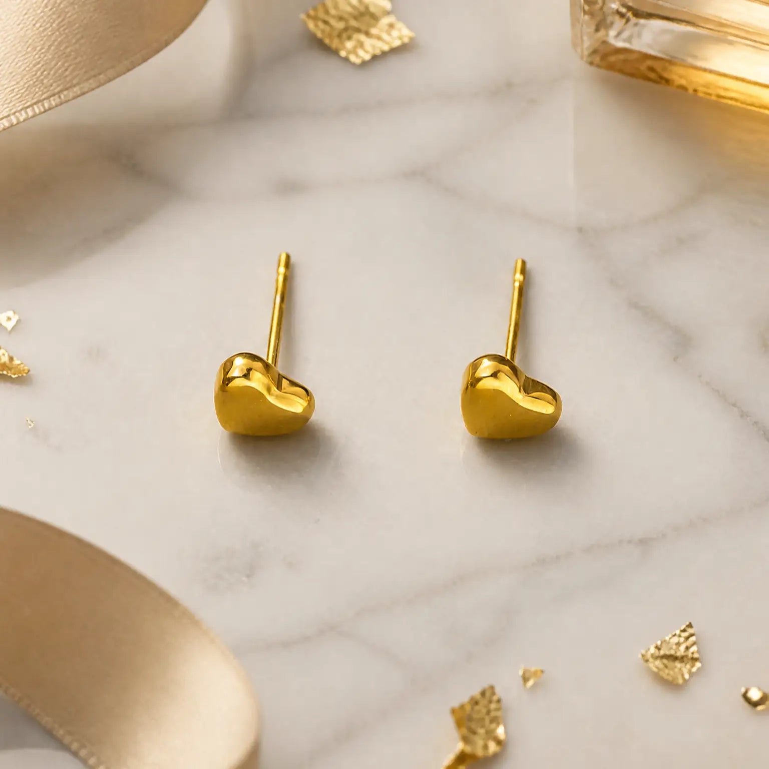 A pair of small gold heart-shaped stud earrings resting on a white marble surface with decorative gold foil pieces and soft beige ribbon props.