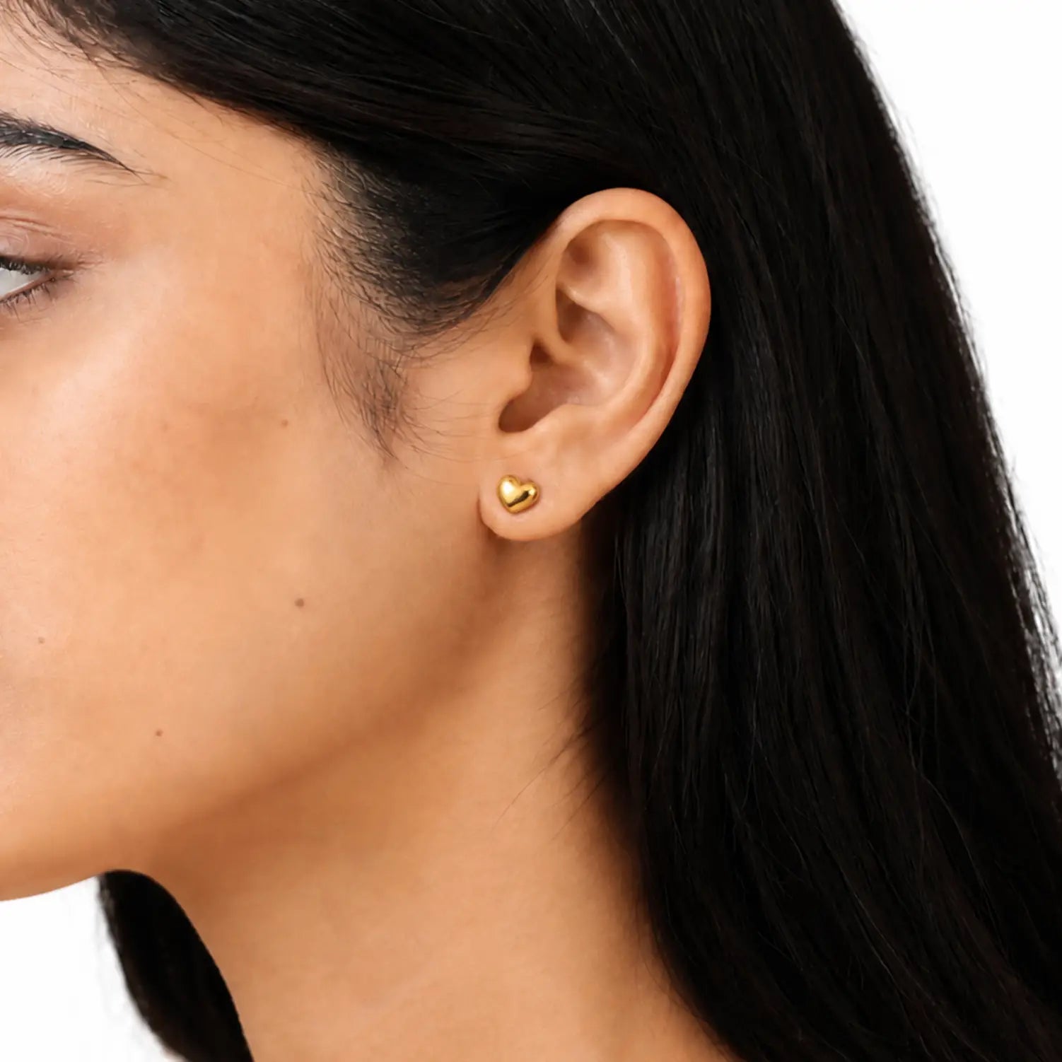 A close-up of a woman's ear wearing a small, polished gold heart-shaped stud earring against a clean white background.