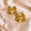 A pair of shiny gold bow-shaped stud earrings displayed on a soft cream satin fabric background with subtle jewelry props at the edges.
