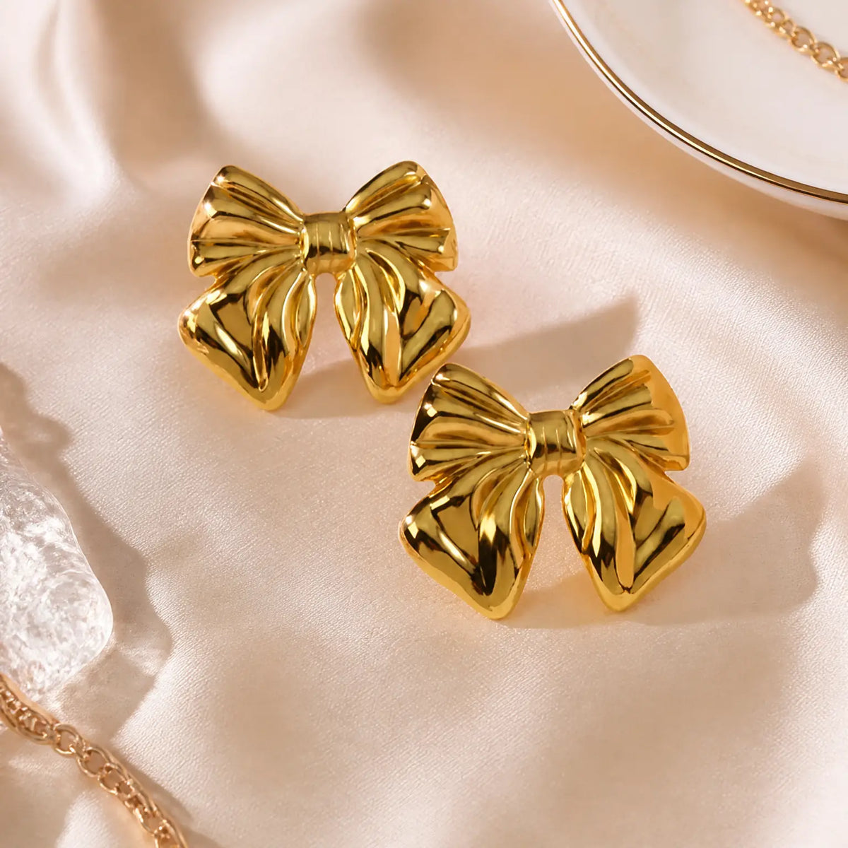 A pair of shiny gold bow-shaped stud earrings displayed on a soft cream satin fabric background with subtle jewelry props at the edges.