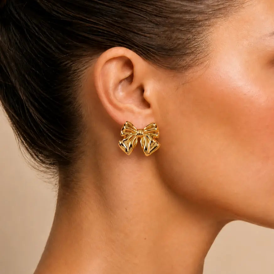 A close-up of a model's ear wearing a small gold bow-shaped stud earring against a soft beige background.
