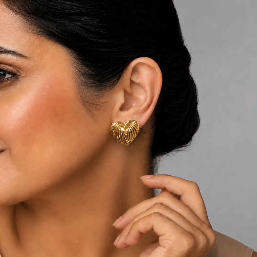 A close-up of a woman wearing a textured gold heart-shaped stud earring, showing the earring on her ear against a neutral gray background.