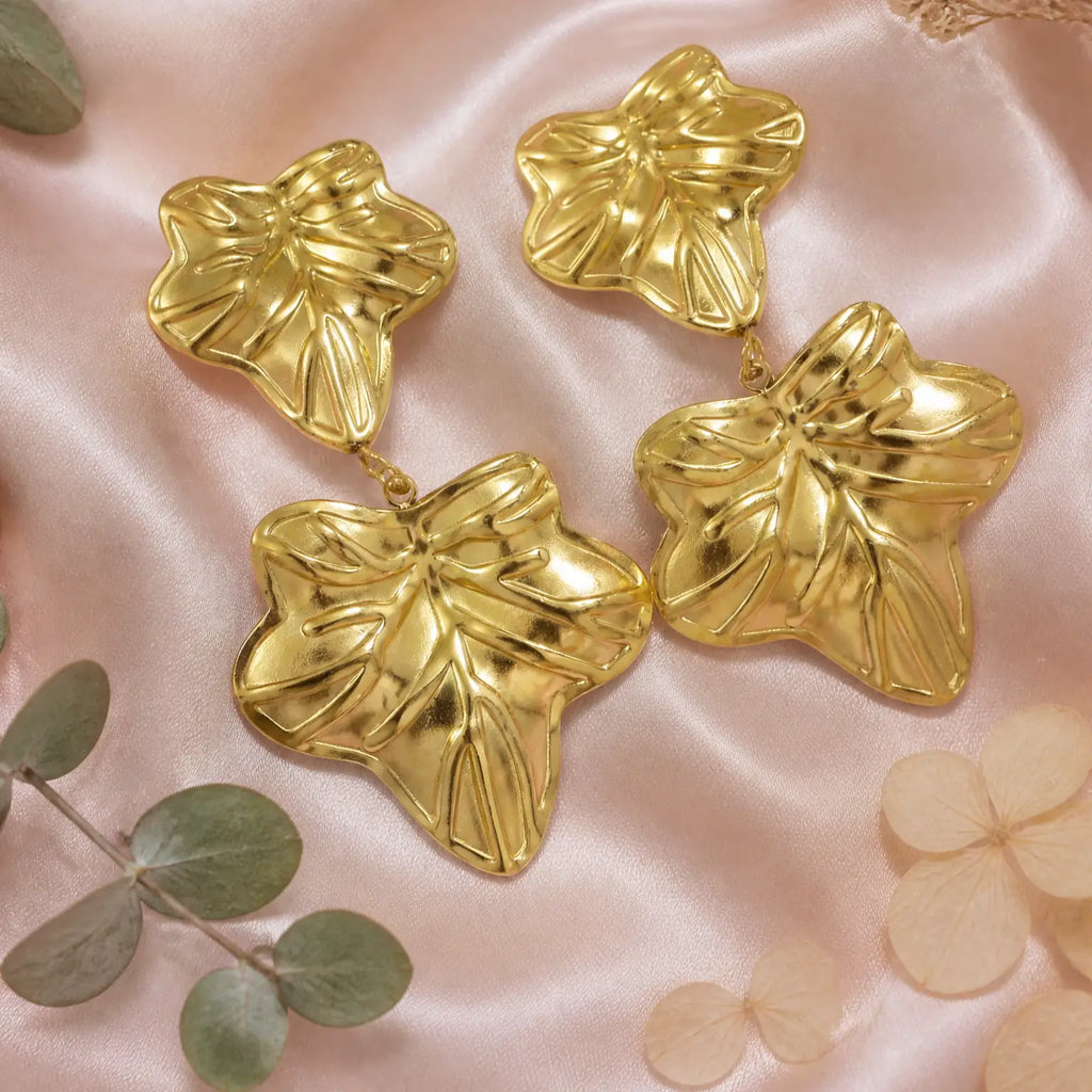 Four shiny gold leaf-shaped earrings arranged on soft pink satin fabric with decorative green leaves and pale petals around them.