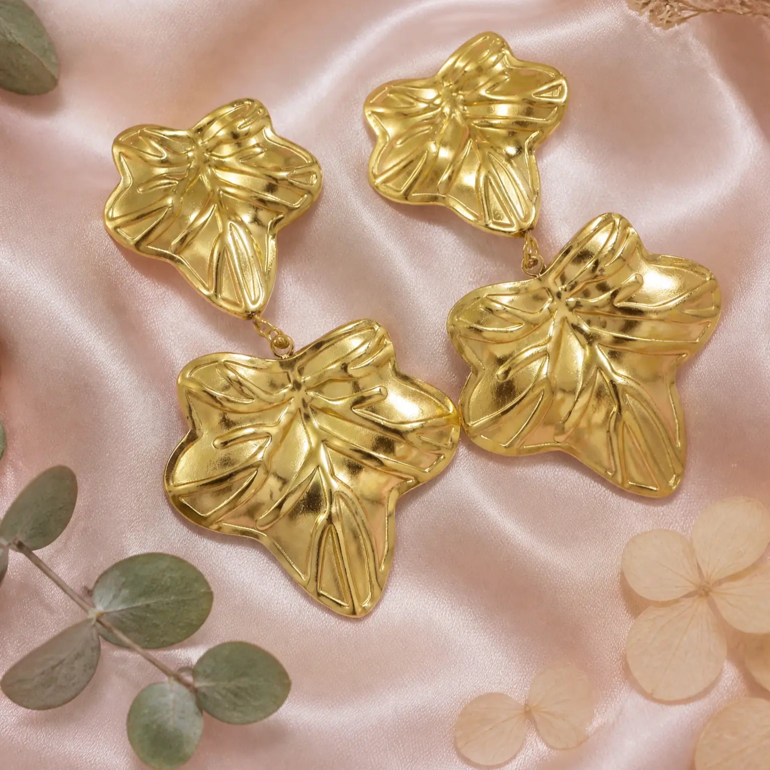 Four shiny gold leaf-shaped earrings arranged on soft pink satin fabric with decorative green leaves and pale petals around them.