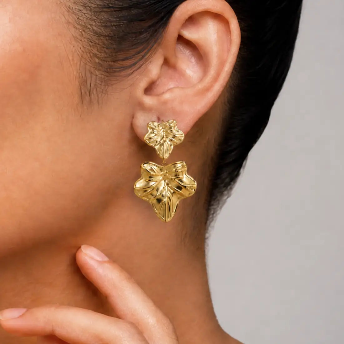 A close-up of a model's ear wearing a pair of textured gold dangle earrings, with her hand resting near her neck against a neutral gray background.