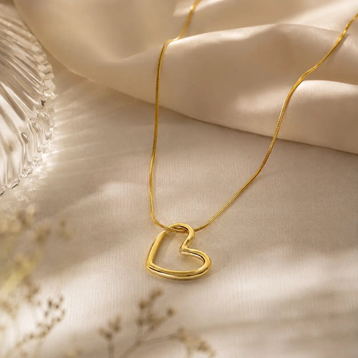 A delicate gold chain necklace with an open heart pendant, styled on draped cream satin fabric with soft shadows.