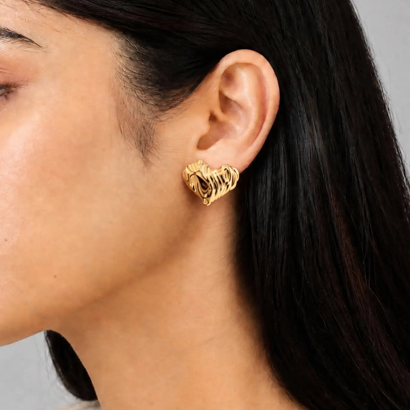 A close-up of a woman's ear wearing a textured gold heart-shaped stud earring.