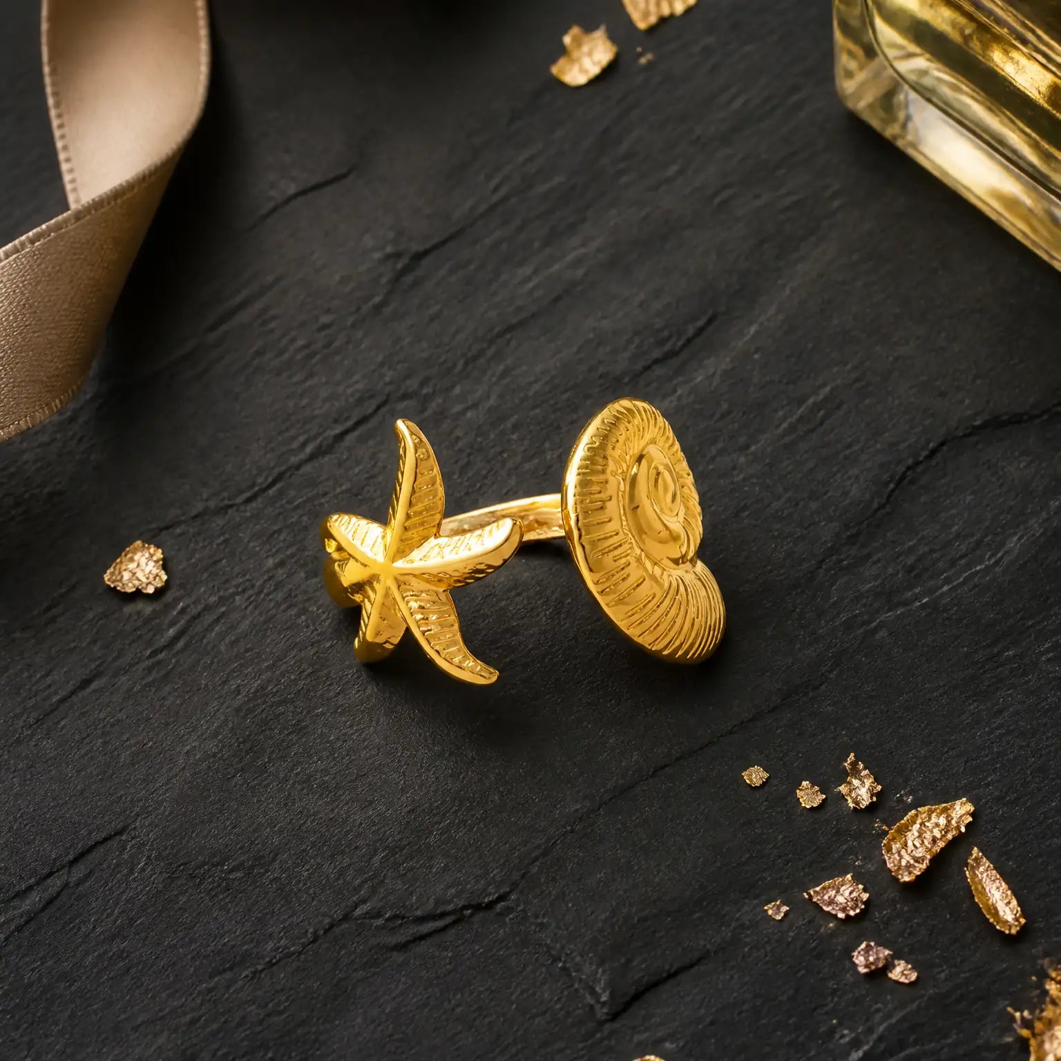 A pair of gold cufflinks shaped like a starfish and a spiral shell, displayed on a dark textured slate surface with scattered gold foil flakes.