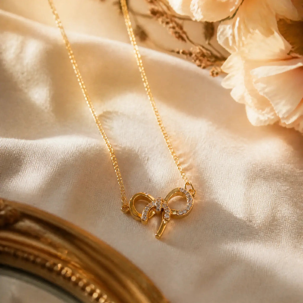 A delicate gold necklace with a bow-shaped pendant adorned with small clear stones, resting on draped cream satin fabric with soft floral accents.