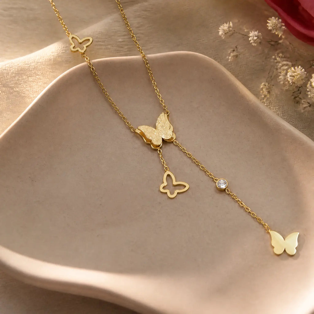 A delicate gold Y-necklace featuring butterfly charms, a small textured butterfly, an outline butterfly, and a clear rhinestone, displayed on a beige ceramic dish.