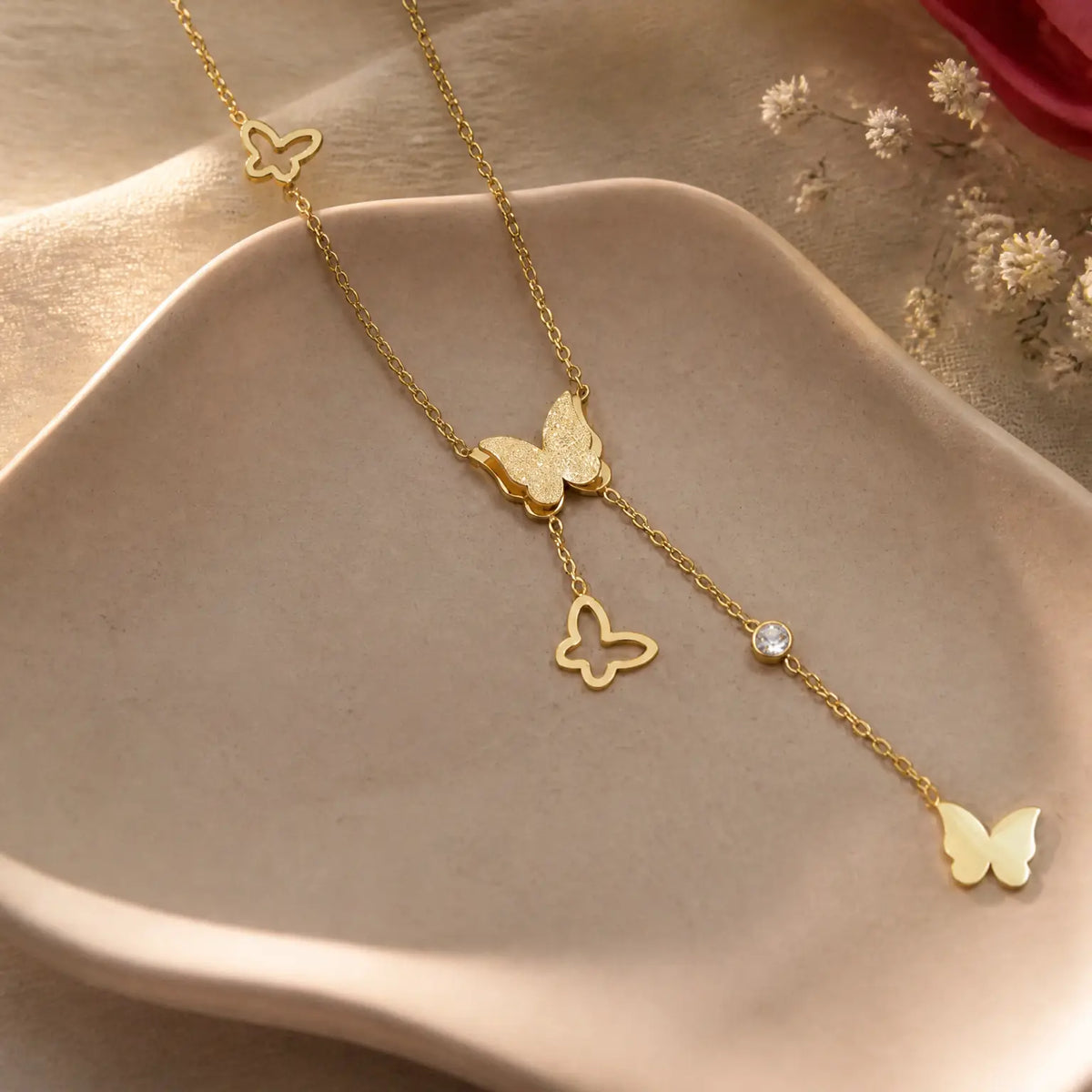 A delicate gold Y-necklace featuring butterfly charms, a small textured butterfly, an outline butterfly, and a clear rhinestone, displayed on a beige ceramic dish.