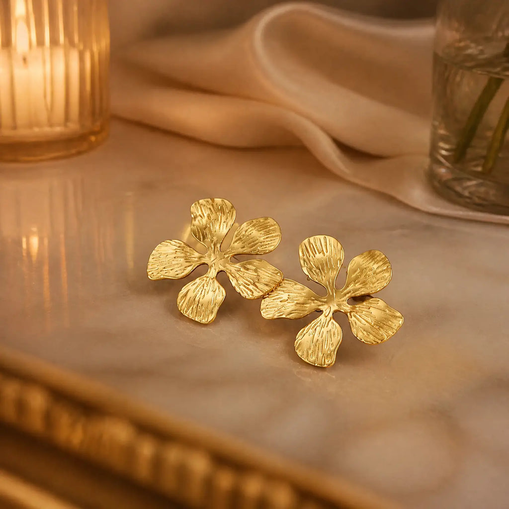 A pair of textured gold flower-shaped earrings resting on a polished marble surface, with a softly blurred warm-toned background featuring draped fabric and a candle.
