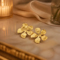 A pair of textured gold flower-shaped earrings resting on a polished marble surface, with a softly blurred warm-toned background featuring draped fabric and a candle.