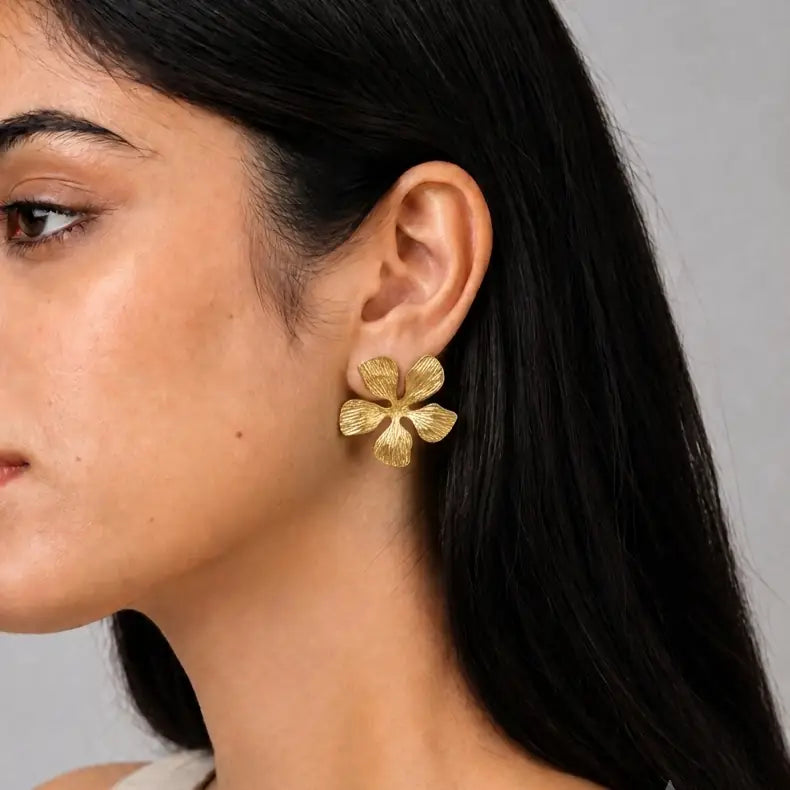 A close-up of a woman's ear wearing a textured gold four-petal flower stud earring against a neutral gray background.