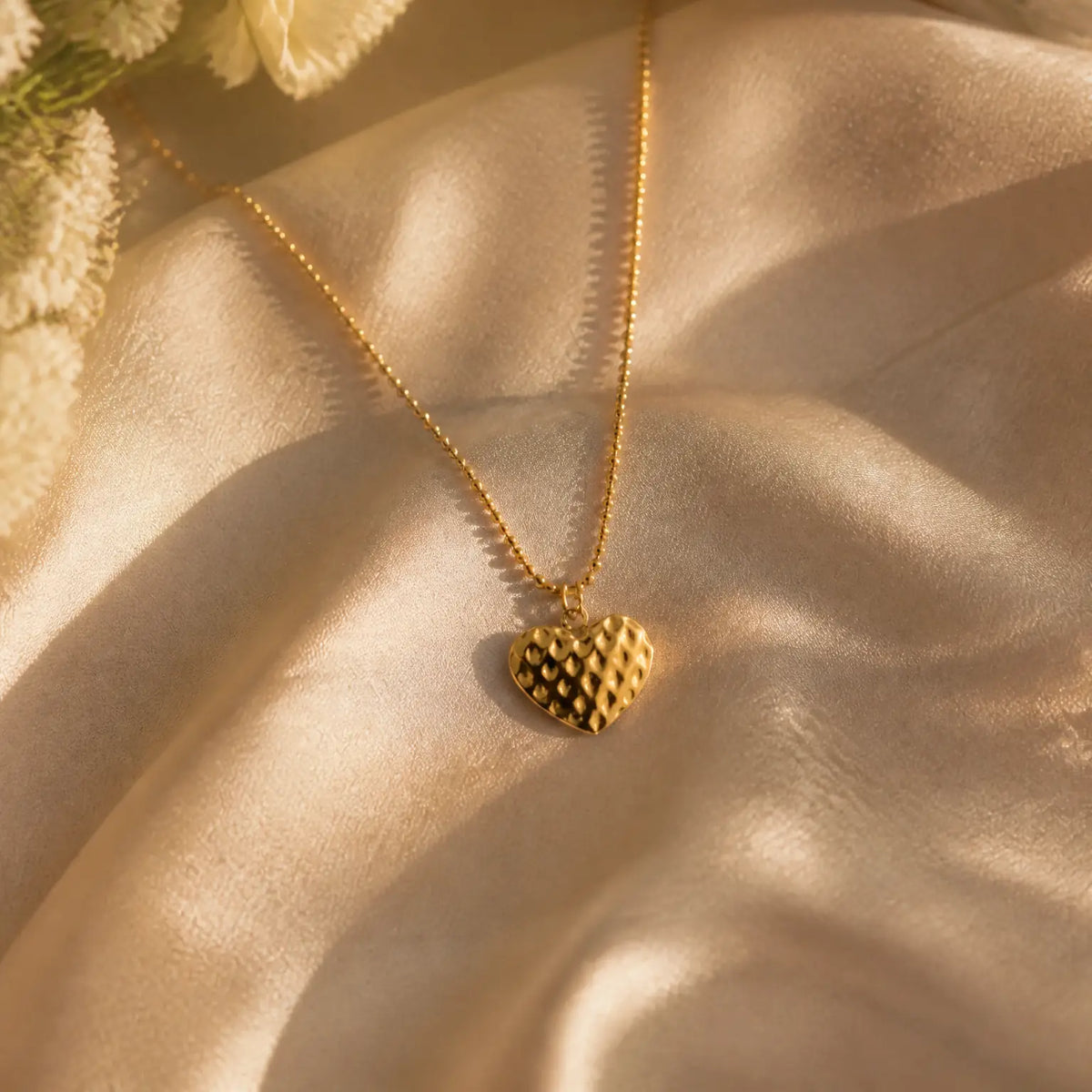 A gold heart pendant necklace with a textured, dimpled surface on a delicate gold chain, displayed on draped champagne-colored satin fabric with soft floral accents.