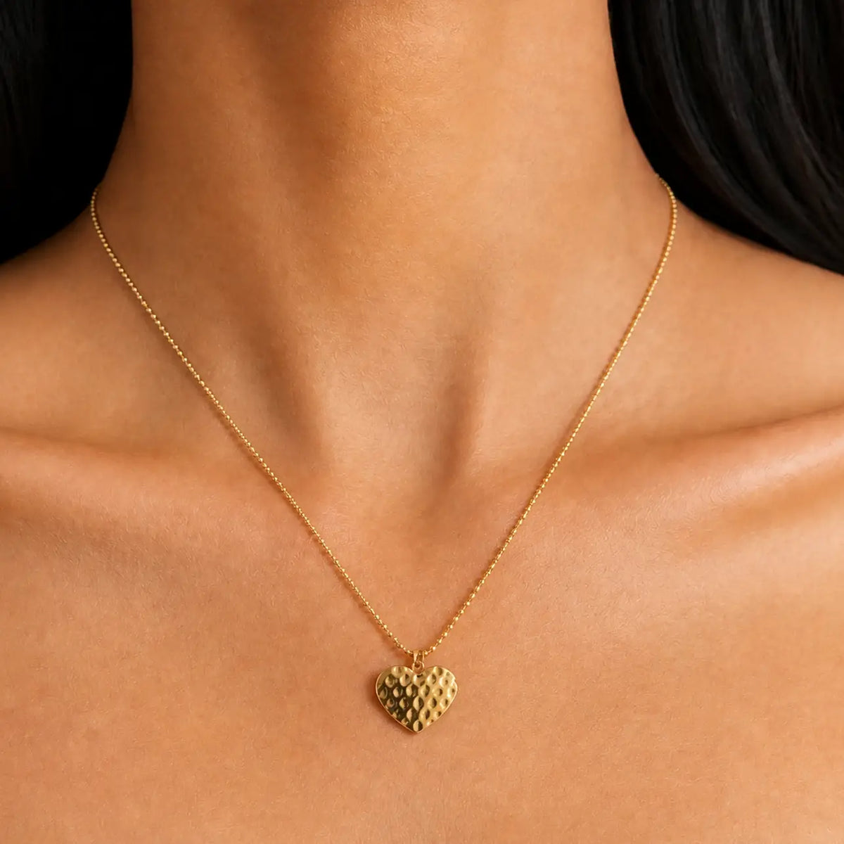 A close-up of a delicate gold chain necklace with a textured heart pendant, worn on a model's neck.