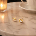 A pair of small gold heart-shaped stud earrings resting on a light marble tabletop, with a softly blurred lit candle on the left and a white cup and saucer on the right.