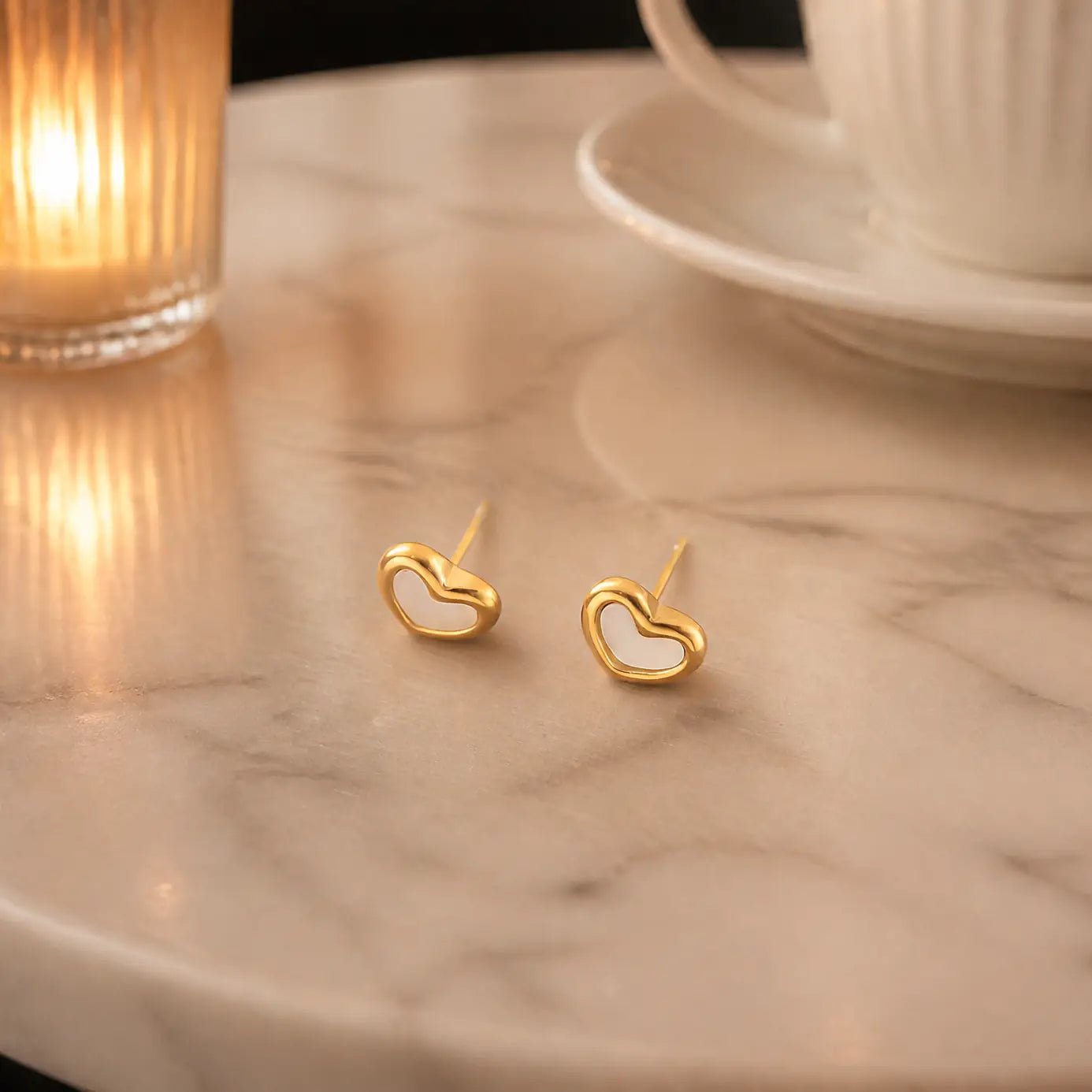 A pair of small gold heart-shaped stud earrings resting on a light marble tabletop, with a softly blurred lit candle on the left and a white cup and saucer on the right.